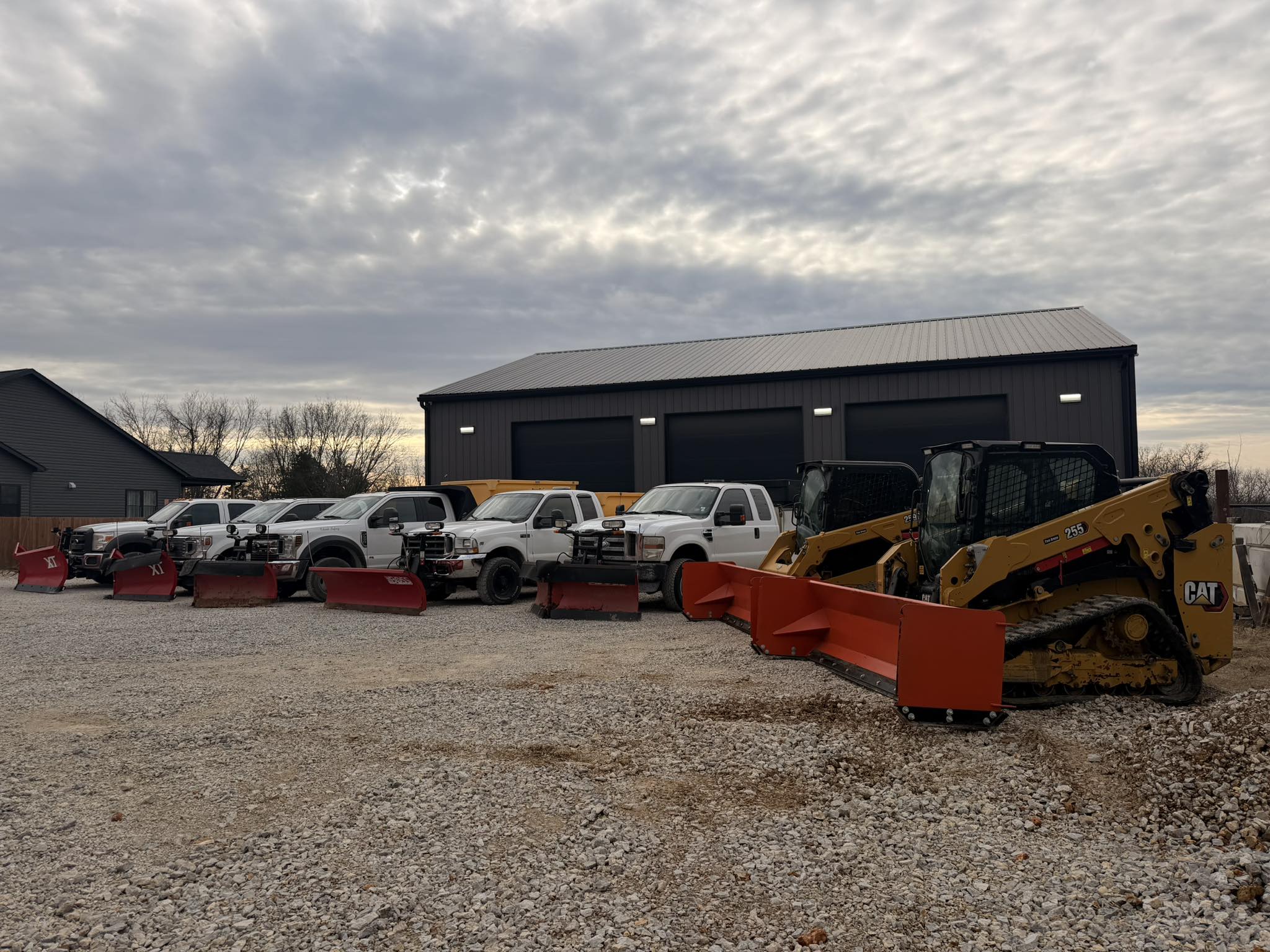 Plow trucks and a skid steer with a snow pusher lined up outside a garage