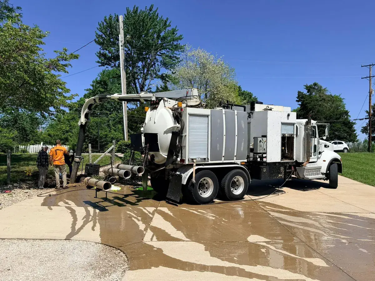 A hydrovac truck at work beside a wet driveway, with two crew members in safety vests