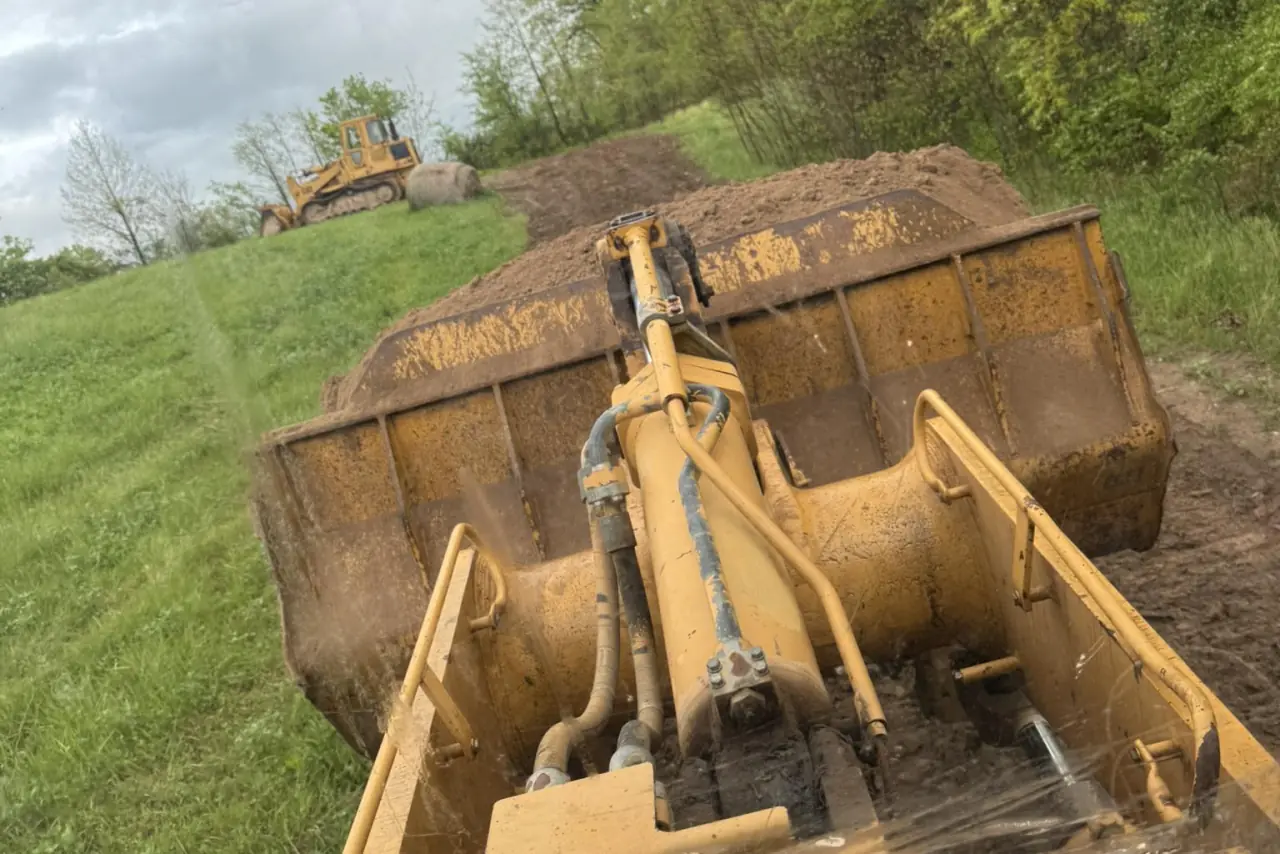 A loader bucket full of dirt on a grassy slope, with a dozer working behind it